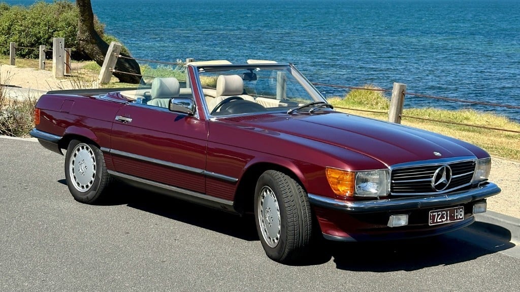 Weinroter Mercedes-Benz SL R107 mit offenem Verdeck an einer Uferstrasse