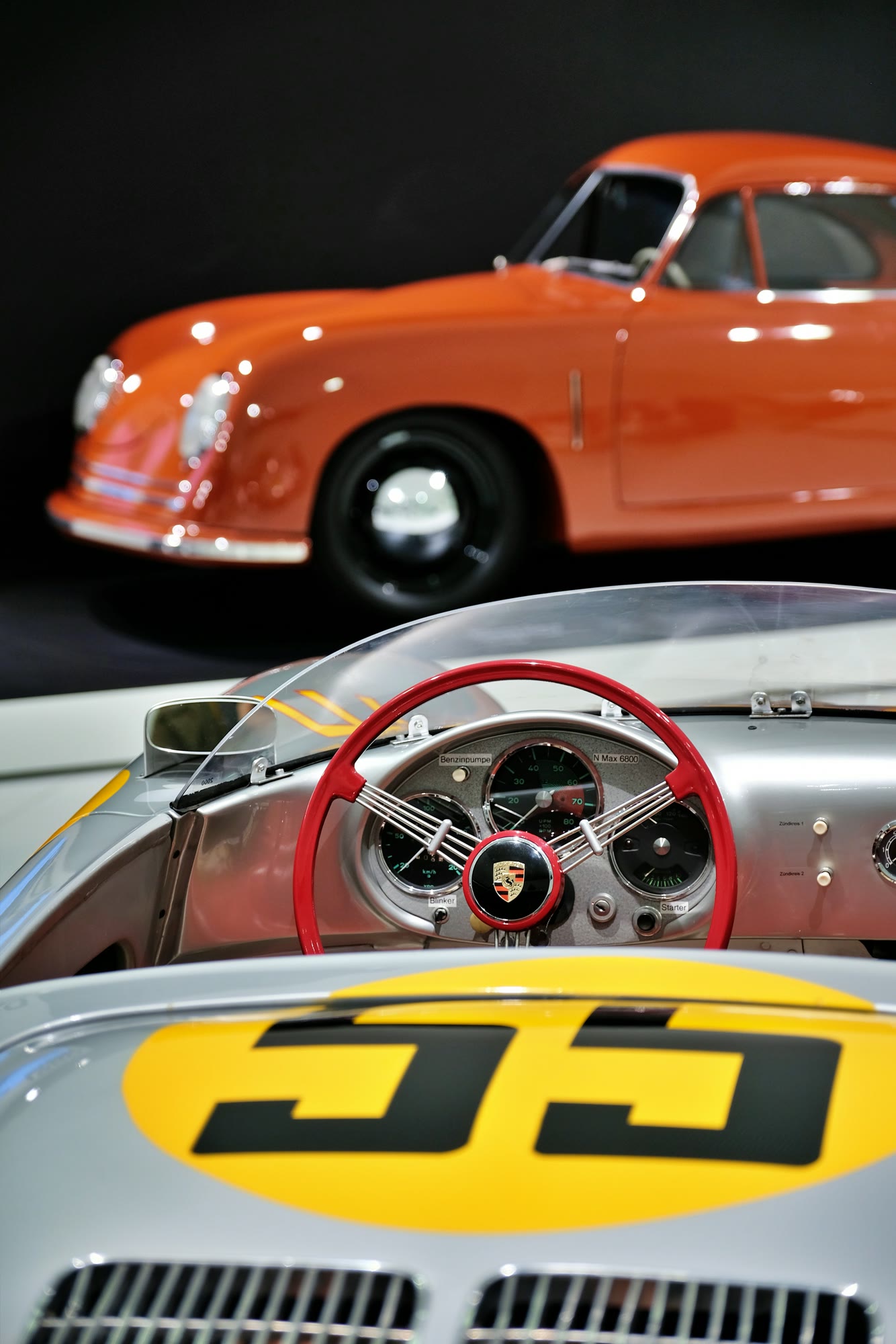 Orange Porsche 356 and silver racing car with number 55 and a red steering wheel